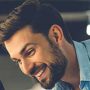 A man with a beard and short hair smiles while looking downwards, wearing a light blue shirt. The blurred background suggests a casual indoor setting reminiscent of peering through the sleek lines of a clearview fence.