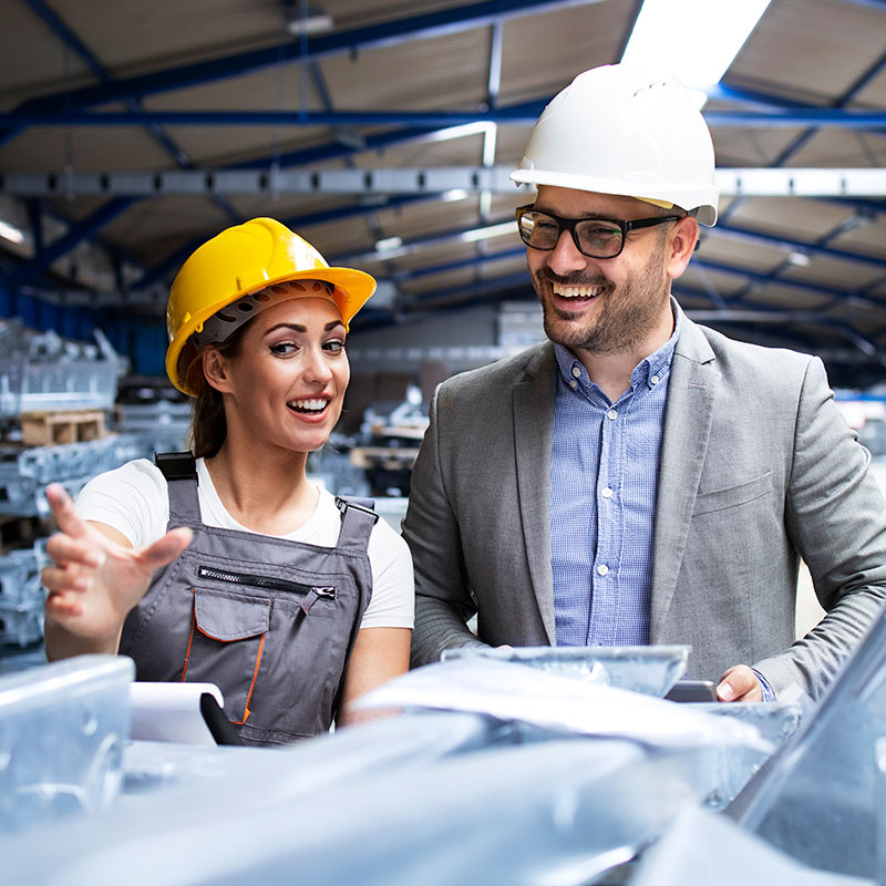 In a bustling industrial setting, two professionals stand equipped with safety helmets. The one on the left in yellow gear gestures and smiles, embodying the spirit of a dedicated manufacturer. Meanwhile, the individual in a white helmet beams, clipboard in hand, ensuring every detail has a clear view.