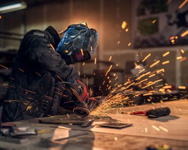 A person wearing a protective welding helmet and gloves is using a power tool to grind metal in a workshop. Sparks fly from the tool, illuminating the space, while various tools and materials are scattered on the wooden workbench behind a clear view fence.