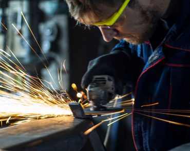 A person wearing safety goggles operates a grinding tool on a piece of metal near a clear view fence, producing bright sparks. The individual is focused, with the blurred industrial background adding depth to their meticulous work.