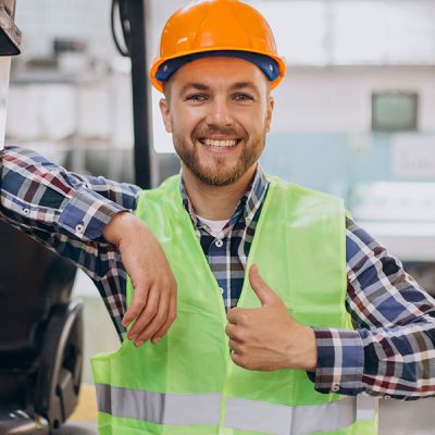 A smiling man in an orange hard hat and neon green safety vest gives a thumbs-up beside towering machinery, with a clear view of the industrial setting. His plaid shirt peeks from beneath the vest, adding a touch of personality to his professional attire.