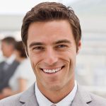 A smiling man in a suit with short brown hair stands indoors, exuding professionalism. The blurred background shows others in business attire, perhaps discussing the latest trends from a leading security fencing manufacturer.