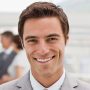 A smiling man in a suit with short brown hair stands indoors, exuding professionalism. The blurred background shows others in business attire, perhaps discussing the latest trends from a leading security fencing manufacturer.