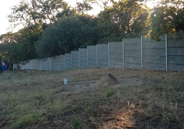 A long, gray ClamberPrufe Clear View Fencing panel runs parallel to a patch of dry, grassy ground with a green bucket and a shovel resting on a dirt mound. Dense trees and shrubs grow behind the fence.