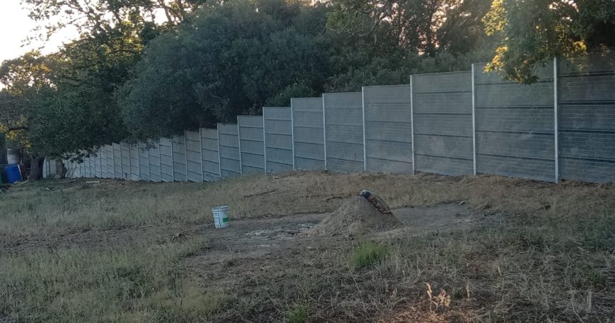 A long, gray ClamberPrufe Clear View Fencing panel runs parallel to a patch of dry, grassy ground with a green bucket and a shovel resting on a dirt mound. Dense trees and shrubs grow behind the fence.