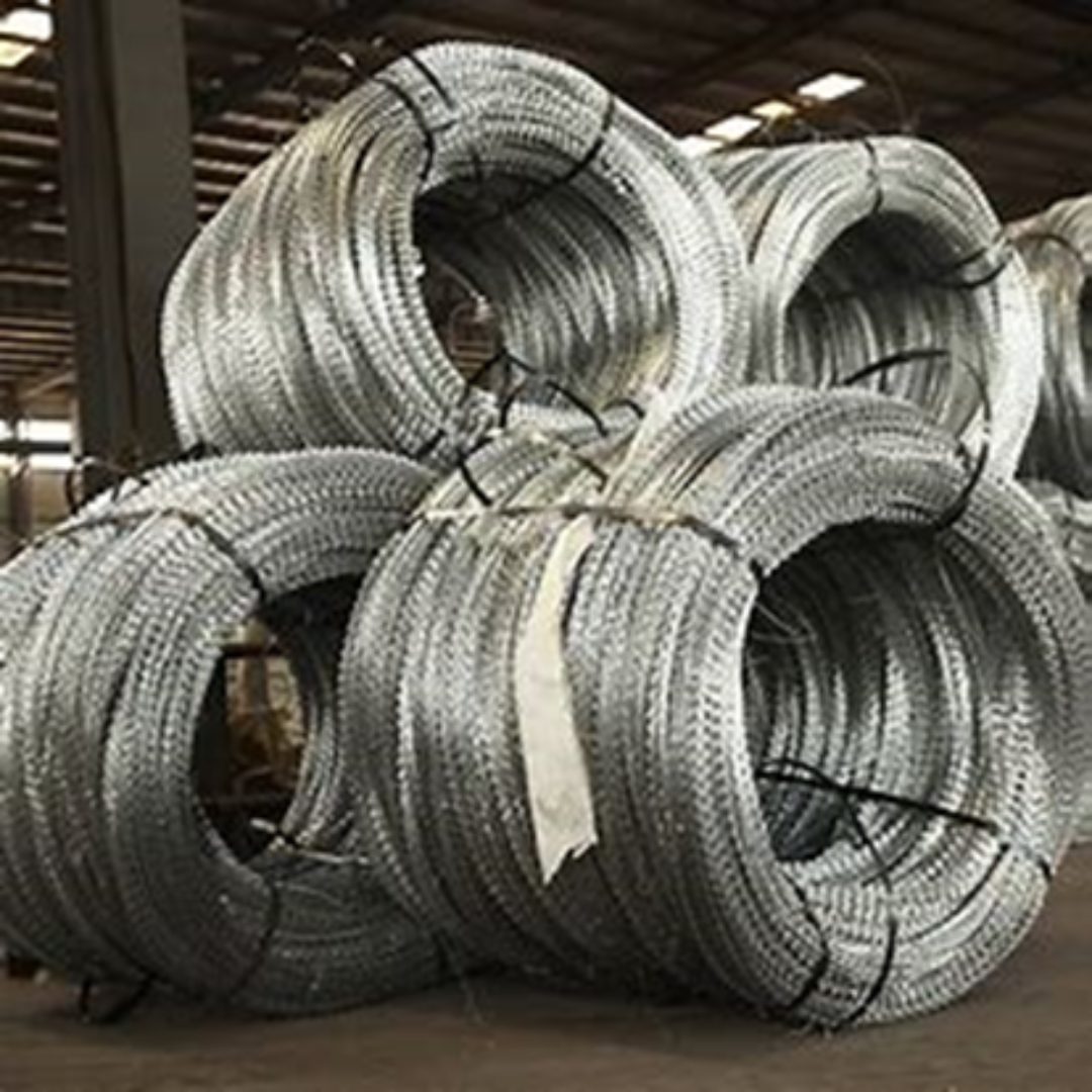 Coils of razor wire are stacked in an industrial warehouse setting by a reputed manufacturer. The metallic coils are tightly wound, with sharp edges visible, affording a clear view of the high ceiling and structural beams typical of Jumalu Fencing products.