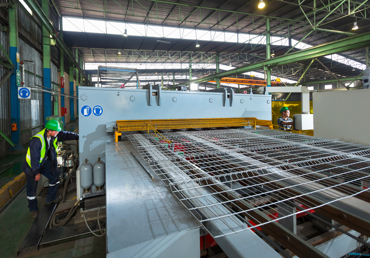 In a factory, workers for a manufacturer operate machinery to process metal grids. The expansive industrial space, under high ceilings and bright overhead lighting, provides a clear view of operations. Safety vests and helmets underscore the commitment to safety and efficient workflow.
