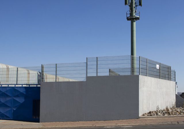 A communication tower stands tall on a fenced platform against the backdrop of a clear blue sky. Surrounded by Jumalu security fencing and a concrete wall at its base, it edges beside a road in an industrial area, with minimal structures or vegetation disrupting the clear view.
