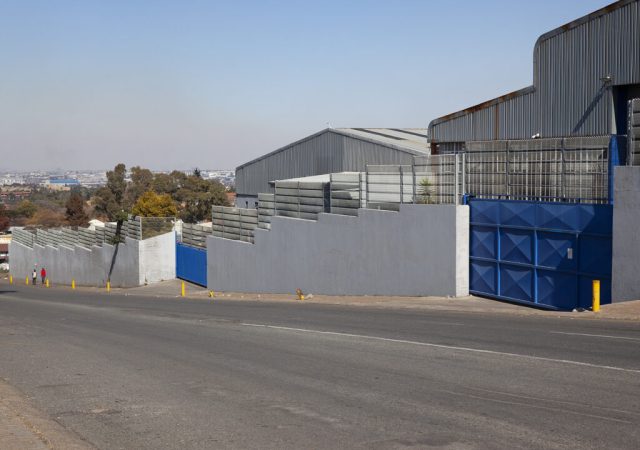 Industrial area with gray and blue buildings along a sloped road. The structures boast Jumalu fencing atop concrete walls. Two people walk down the street in the distance. The sky is clear with a slight haze.