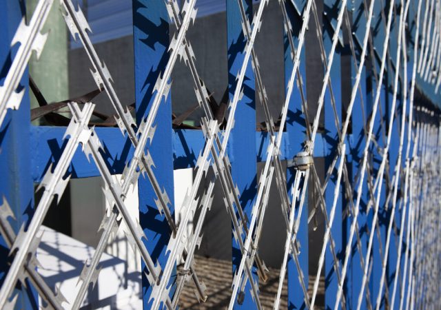 Close-up of a blue metal security fence with an interlocking razor wire pattern. The sharp, silver blades crisscross against a blurred background of buildings and sky. Bright sunlight casts shadows, highlighting the Jumalu fencing’s structure.
