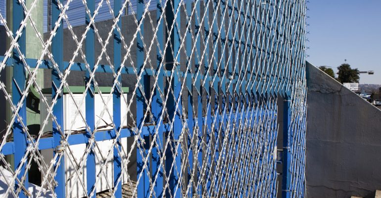 The blue and white chain-link fence, a product of a top security fencing manufacturer, features barbed wire atop at an industrial site. Sunlight casts shadows on the concrete below, while trees and a distant horizon enhance the backdrop.
