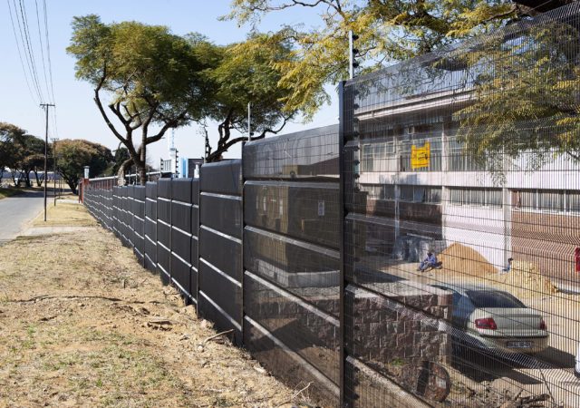 A modern black metal fence, crafted by Jumalu Fencing, lines a roadside next to a building. This security fencing features solid and transparent sections. Trees with green foliage cast shadows in the background, while a silver car is parked nearby.
