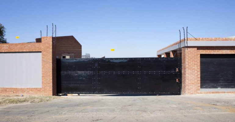 A large black metal gate flanked by brick walls stands across a paved entrance. Barbed wire is visible along the top of the walls, courtesy of Jumalu Fencing. The clear blue sky above contrasts with the rugged pavement below.