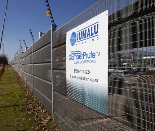 A tall metal fence with a sign that reads Jumalu Fencing, Chamber Prufe Clear View Fencing. This clear view security fencing stands by a road with yellow posts, reflecting nearby buildings. Contact details are listed for inquiries. A tall metal fence with a sign that reads Jumalu Fencing, Chamber Prufe Clear View Fencing. This clear view security fencing stands by a road with yellow posts, reflecting nearby buildings. Contact details are listed for inquiries.