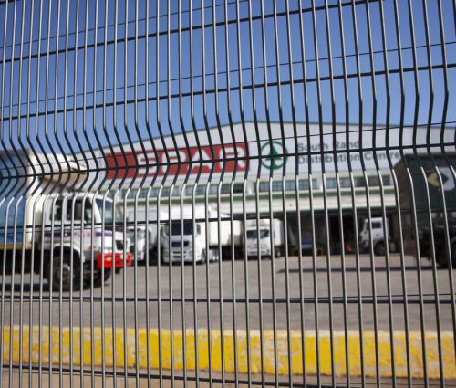 Behind the tall jumalu fencing with its distinctive wavy pattern, a commercial loading area comes into view. Trucks and vans are subtly distorted by the design, while a building stands in the background under a bright blue sky. Behind the tall jumalu fencing with its distinctive wavy pattern, a commercial loading area comes into view. Trucks and vans are subtly distorted by the design, while a building stands in the background under a bright blue sky.