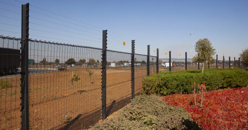 A clear view fence stretches along a dry, brown field dotted with sparse vegetation. Red and green shrubs mark the foreground, while trees and a road with passing vehicles are visible in the background under a bright blue sky.
