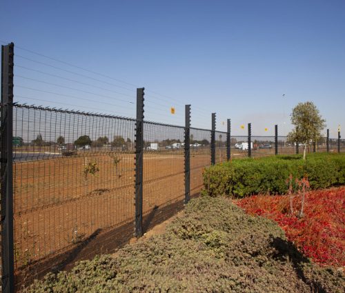 A clear view fence stretches alongside a road with a barren field on one side and a landscaped area featuring green and red foliage offering a clear view on the other. Trees and vehicles can be seen in the background under the pristine blue sky. A clear view fence stretches alongside a road with a barren field on one side and a landscaped area featuring green and red foliage offering a clear view on the other. Trees and vehicles can be seen in the background under the pristine blue sky.