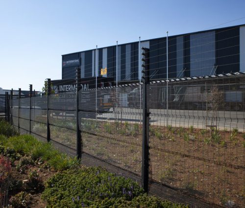 A logistics or warehouse facility with a clearview black fence in front, set against a clear blue sky. The building boasts a modern design and features a sign that reads INTERMODAL, while lush vegetation flourishes in the foreground.