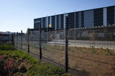 A modern industrial building with a sign reading INTERMODAL stands behind a tall, manufacturer-grade clear view fence topped with barbed wire. The foreground features a garden bursting with various plants and flowers.