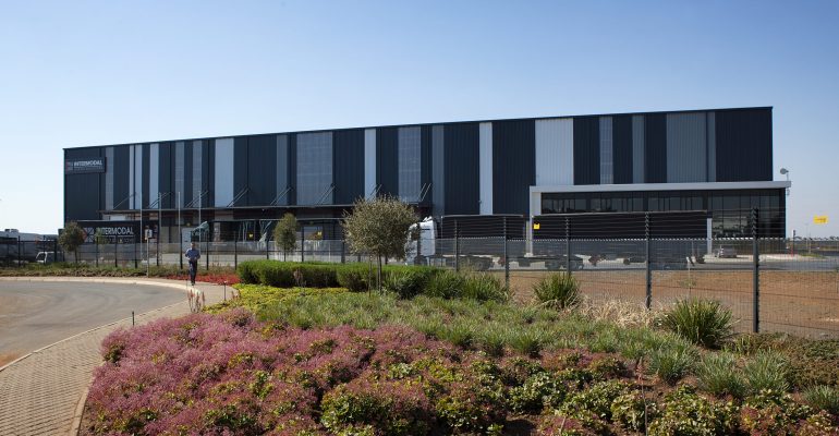 A large, modern warehouse building with a dark gray facade features vertical patterns. In the foreground, pink flowering plants and green shrubs border the Jumalu fencing. The area has a clear view under a bright blue sky.