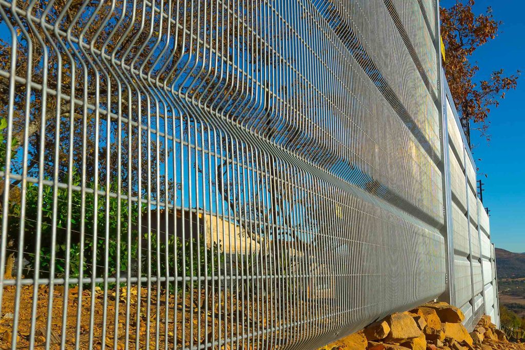Close-up of a ClearView fence with vertical and horizontal bars, casting shadows on a sunny day. The manufacturer designed it with rocks at the base and a tree in the background, all set against a clear blue sky.
