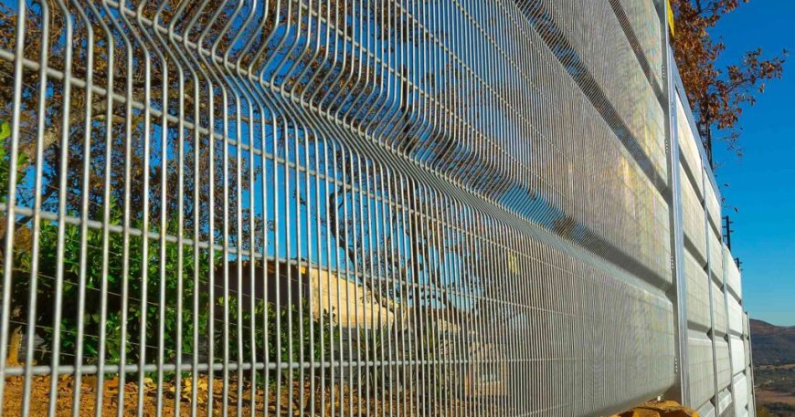A tall metal security fence with closely spaced vertical bars stretches into the distance, set against a bright blue sky. The fence runs alongside a rocky, sloped terrain with sparse vegetation and bare trees. Its design ensures a clear view of the landscape beyond.