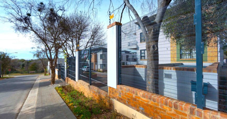 Street view of a residential area with a brick and metal fence bordering a modern house. Leafless trees line the sidewalk next to the paved road under a clear blue sky, offering a clear view. A yellow sign is mounted on the fence.